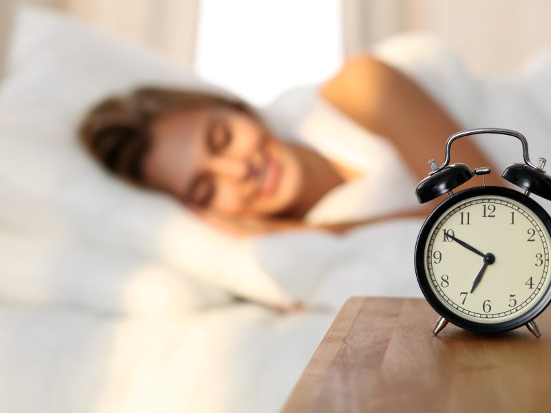 Beautiful young woman sleeping and smiling while lying in bed comfortably and blissfully on the background of alarm clock is going to ring. Sunbeam dawn  on her face.