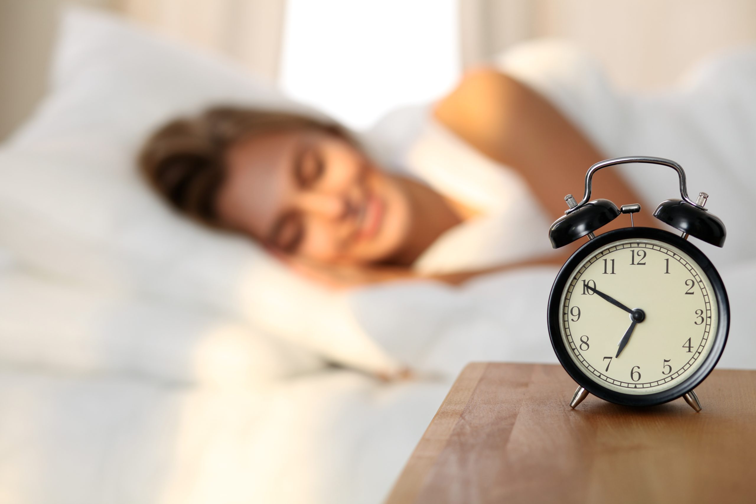 Beautiful young woman sleeping and smiling while lying in bed comfortably and blissfully on the background of alarm clock is going to ring. Sunbeam dawn  on her face.
